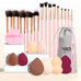 Set of 25 professional makeup tools with pink-handled brushes, sponge balls, powder puffs, eyebrow trimmer, and plush storage bag.